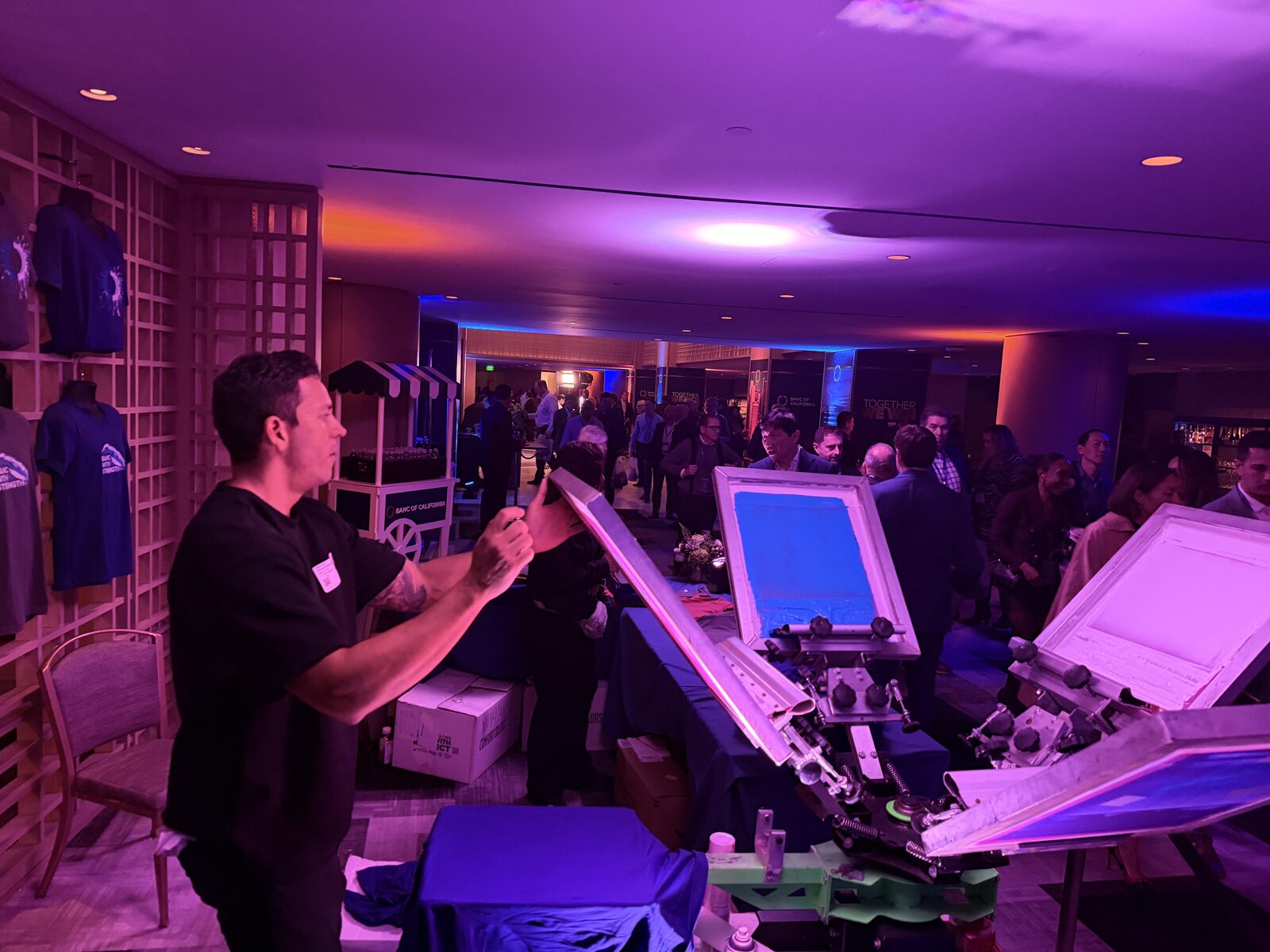 Printer raising a screen on a four-color press while suited guests mingle at a purple-lit corporate gala