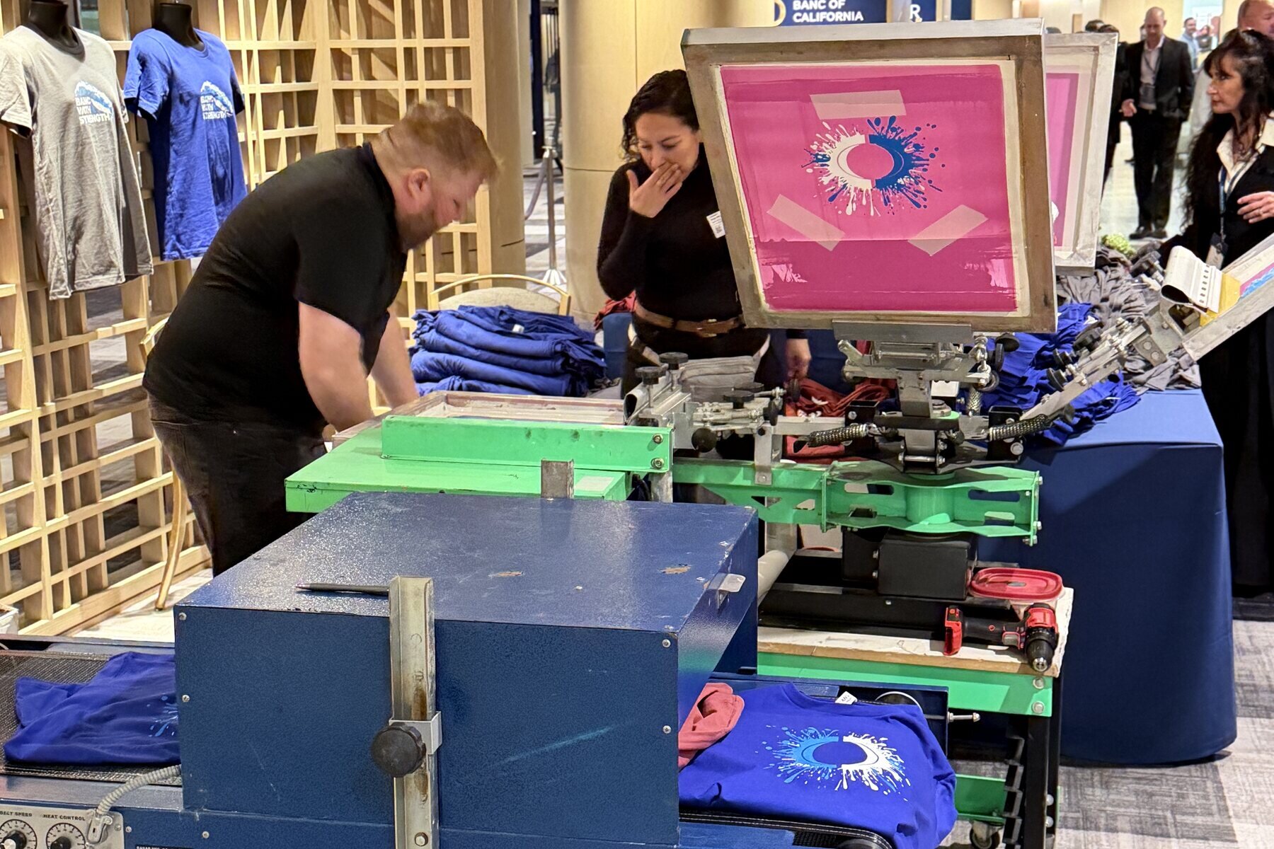 Printers pulling a bright pink screen print onto shirts beside stacks of blue tees