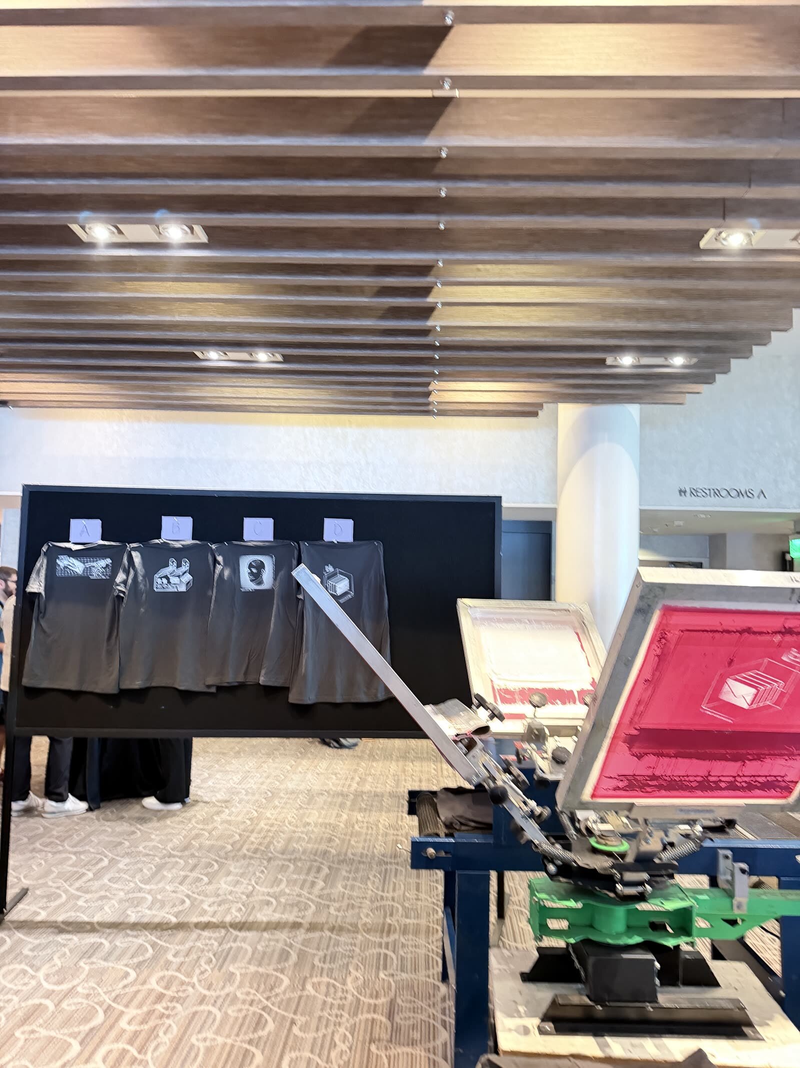 Screen-printing press beside a display wall of finished black shirts in a venue corridor