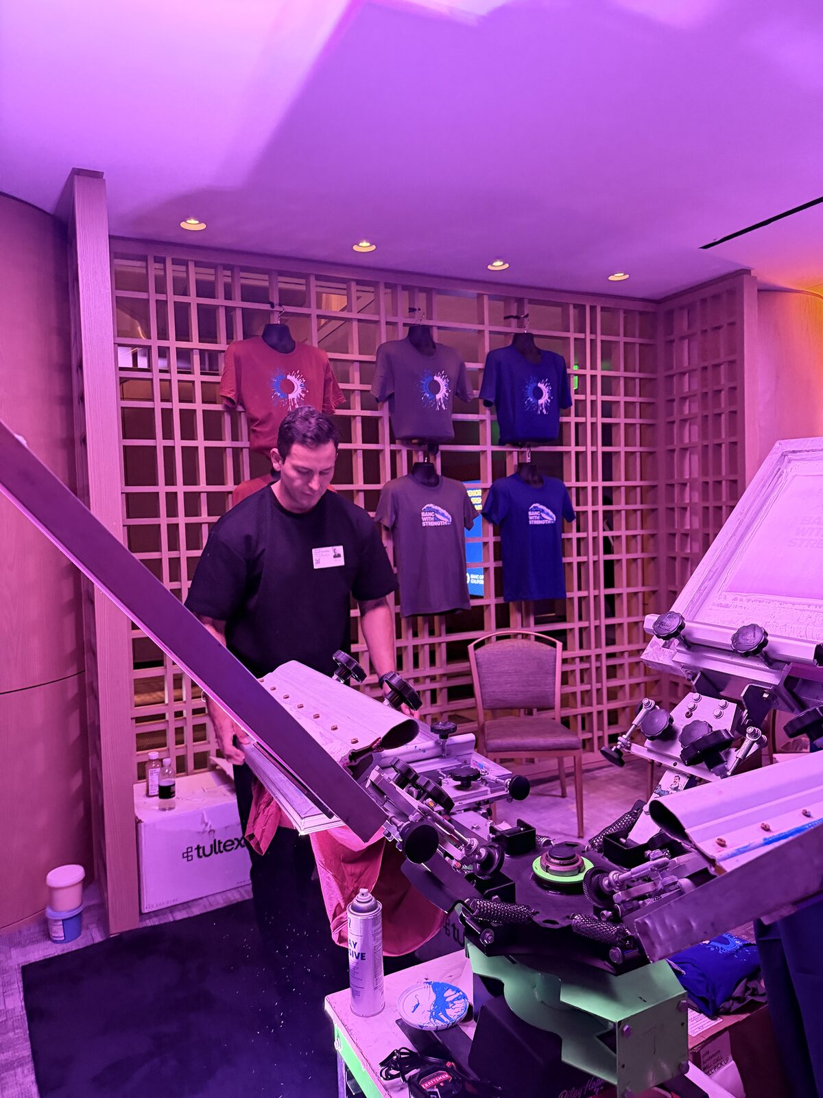 Printer loading a red shirt onto a press pallet in front of a lattice wall of printed tees