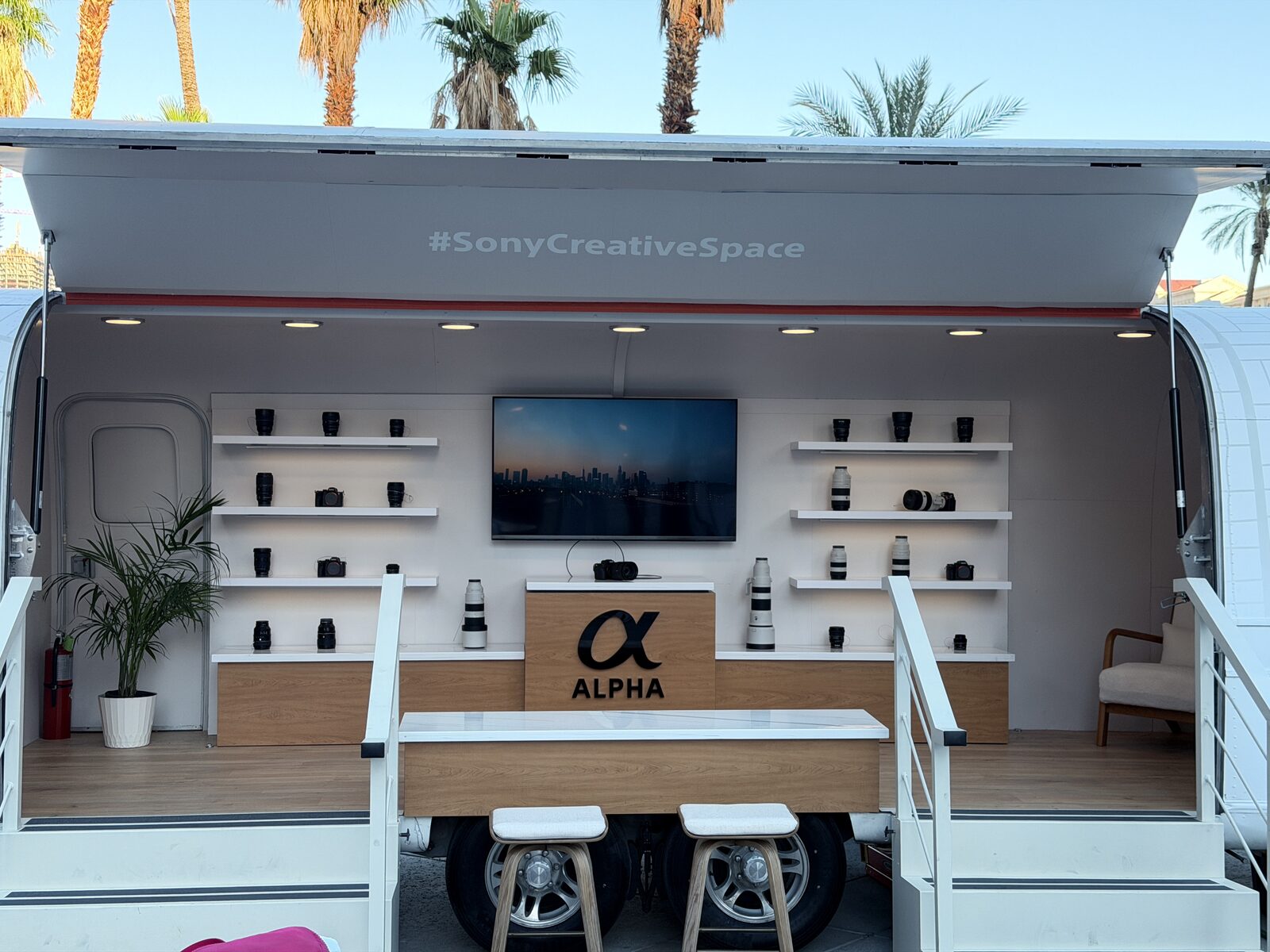 Open-sided event trailer booth with shelf displays and a video screen under palm trees