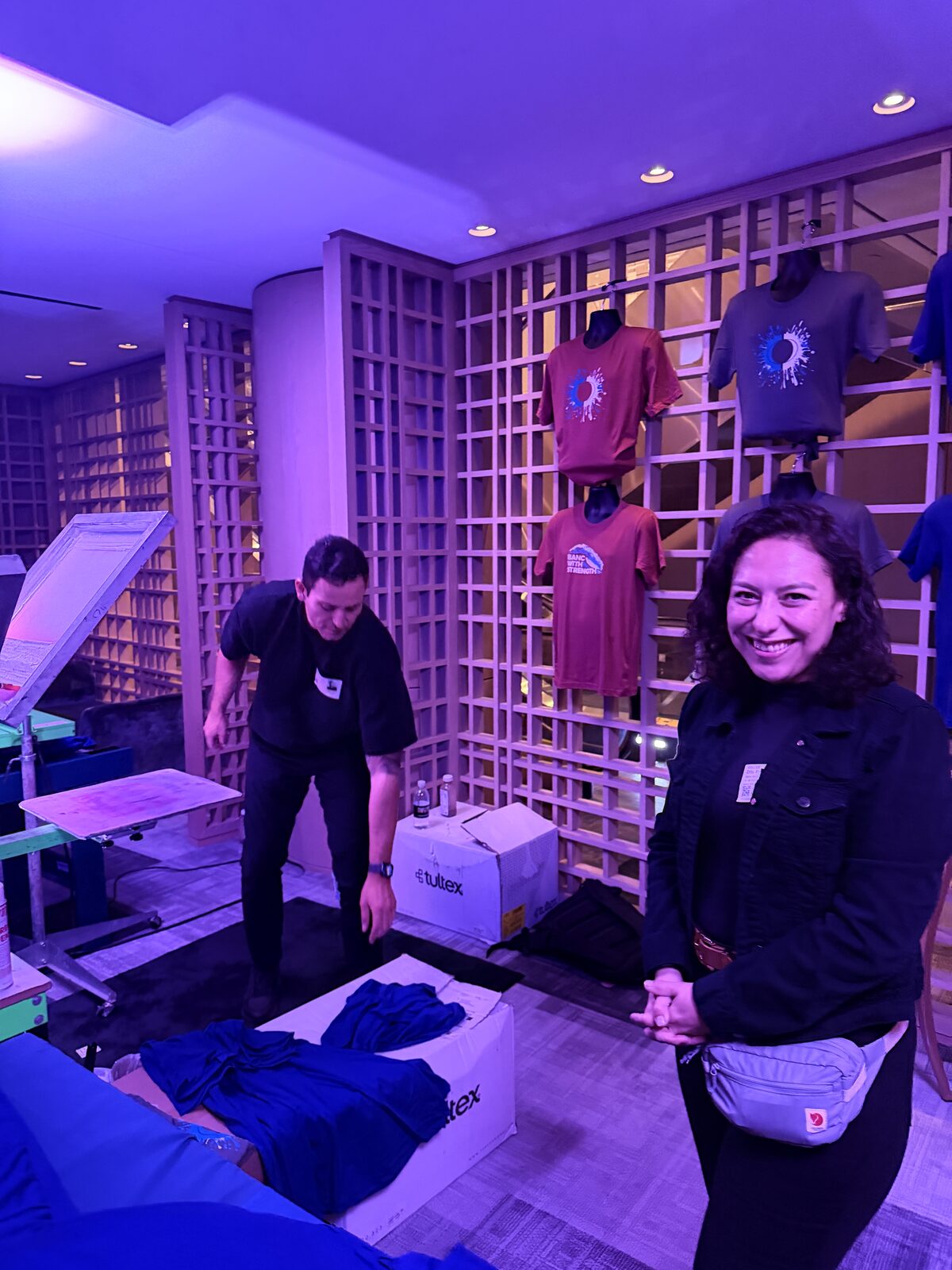 Print crew member smiling at a purple-lit event station with a shirt display wall and garment boxes